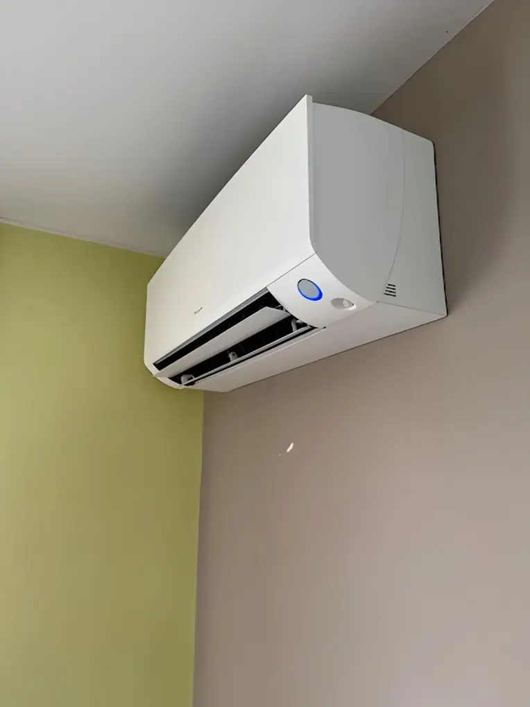 Climatiseur mural blanc vu en contre-plongée, posé haut sur un mur beige à côté d’un mur vert pâle