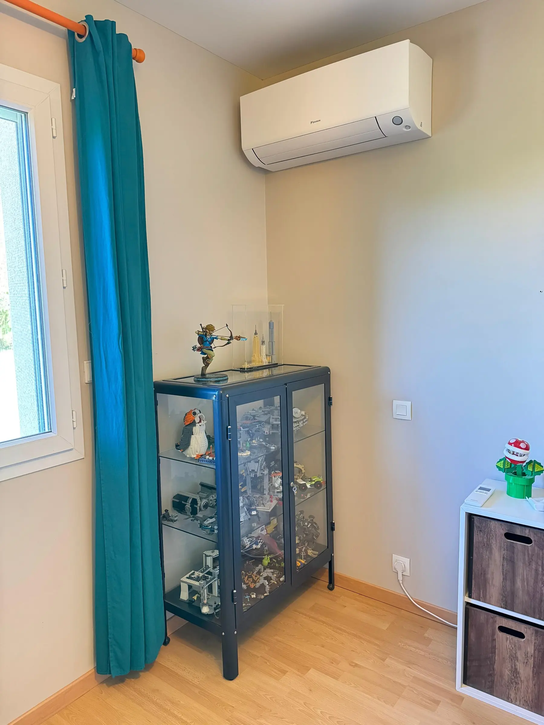 Climatiseur mural blanc fixé en haut d’un mur crème, à côté d’une fenêtre au rideau bleu canard, une vitrine à figurines en dessous