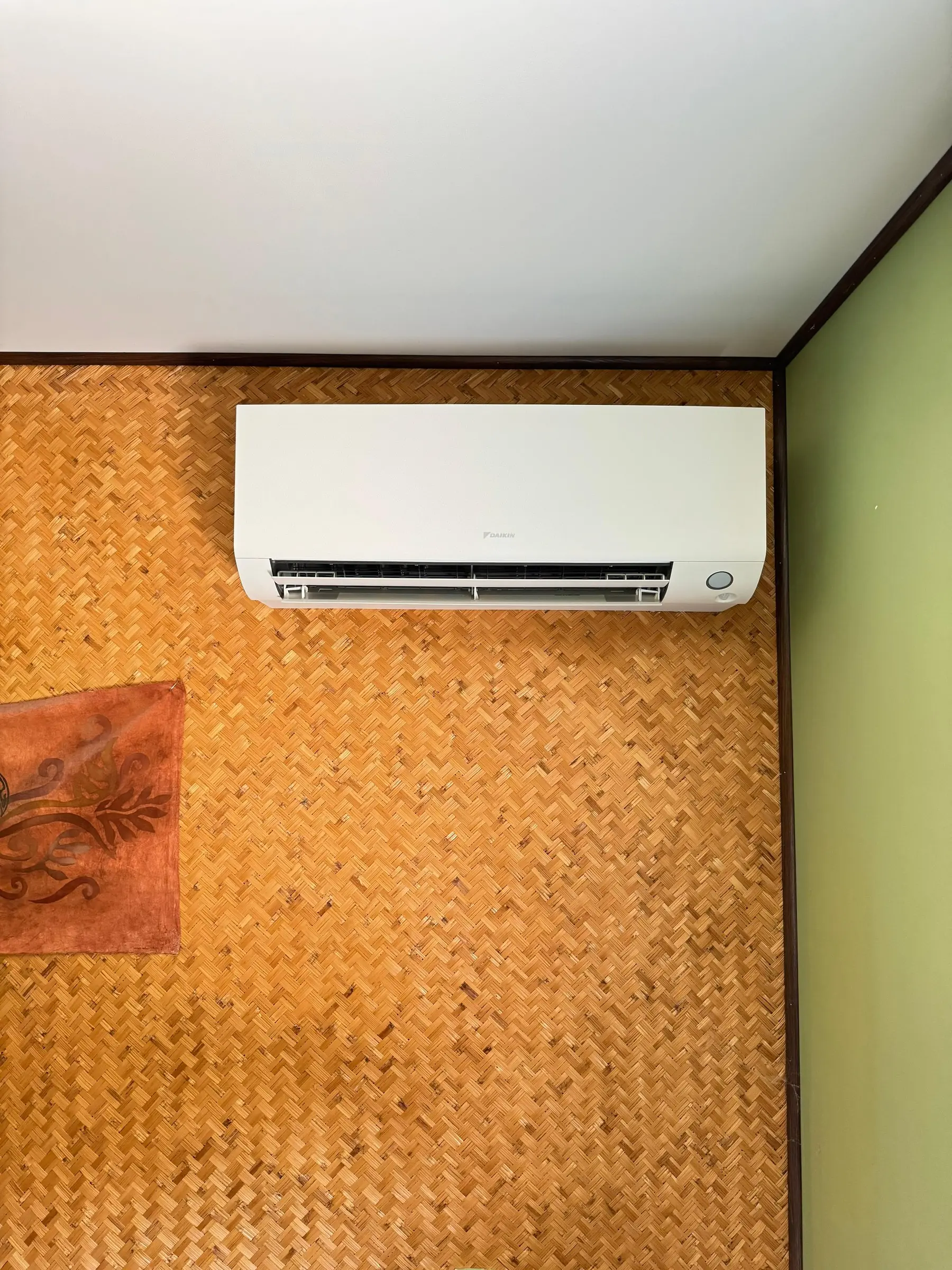 Climatiseur mural blanc posé juste sous un plafond blanc, sur un mur revêtu de paille tressée orangée, à côté d’un mur vert