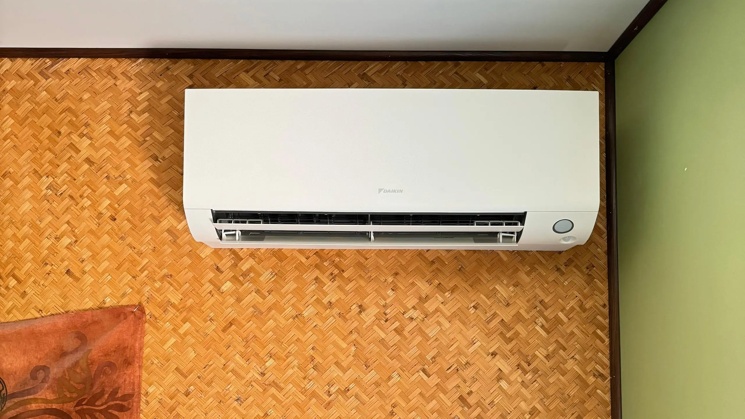 Climatiseur mural blanc posé juste sous un plafond blanc, sur un mur revêtu de paille tressée orangée, à côté d’un mur vert