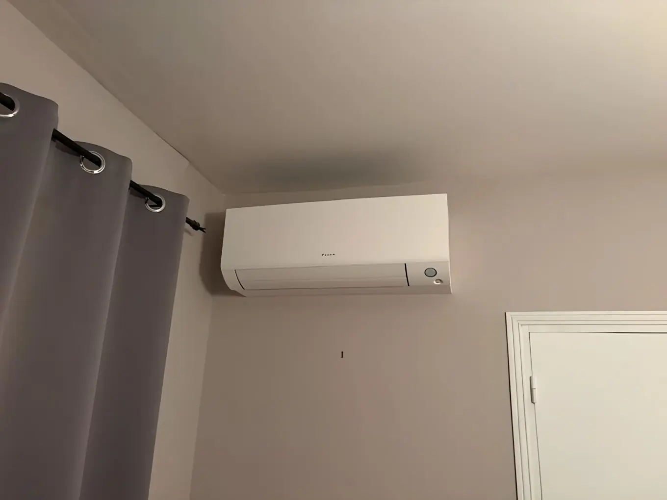 Climatiseur mural blanc en haut d’un mur beige, rideau gris à gauche, porte blanche à droite