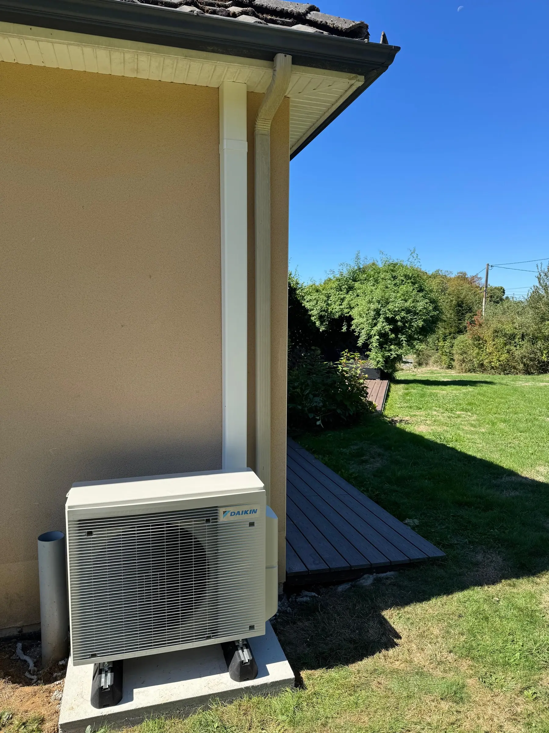 Groupe extérieur de climatisation blanc sur un socle à pieds, au pied d’un mur crème, à côté d’une terrasse en bois et d’une pelouse au soleil