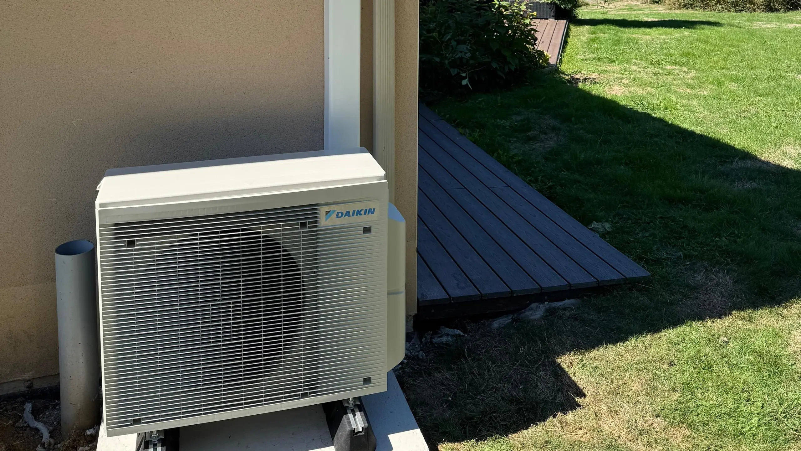 Groupe extérieur de climatisation blanc sur un socle à pieds, au pied d’un mur crème, à côté d’une terrasse en bois et d’une pelouse au soleil