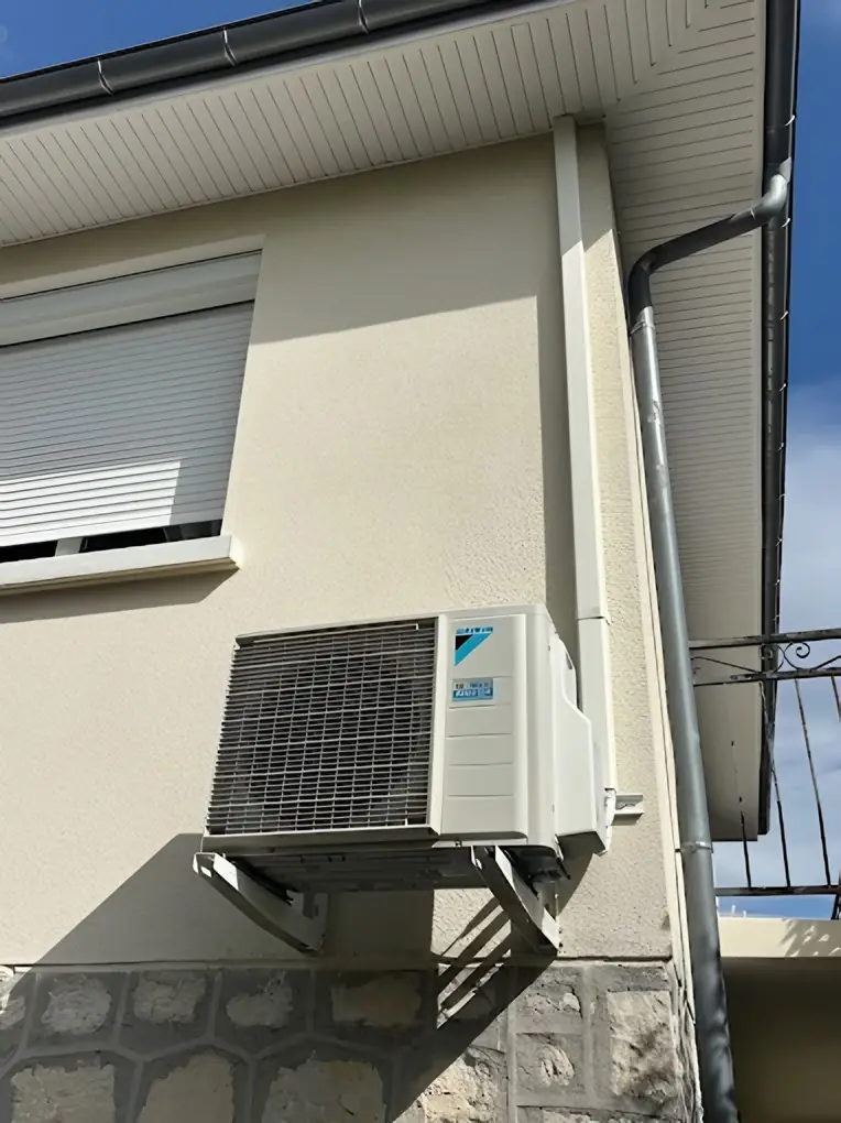Groupe extérieur de climatisation fixé sur des équerres au mur crème d’une maison, sous une fenêtre à volet roulant, soubassement en pierre