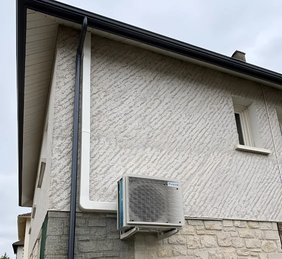 Groupe extérieur de climatisation sur équerres contre une façade au crépi blanc, goulotte blanche jusqu’au toit, descente d’eau et soubassement en pierre