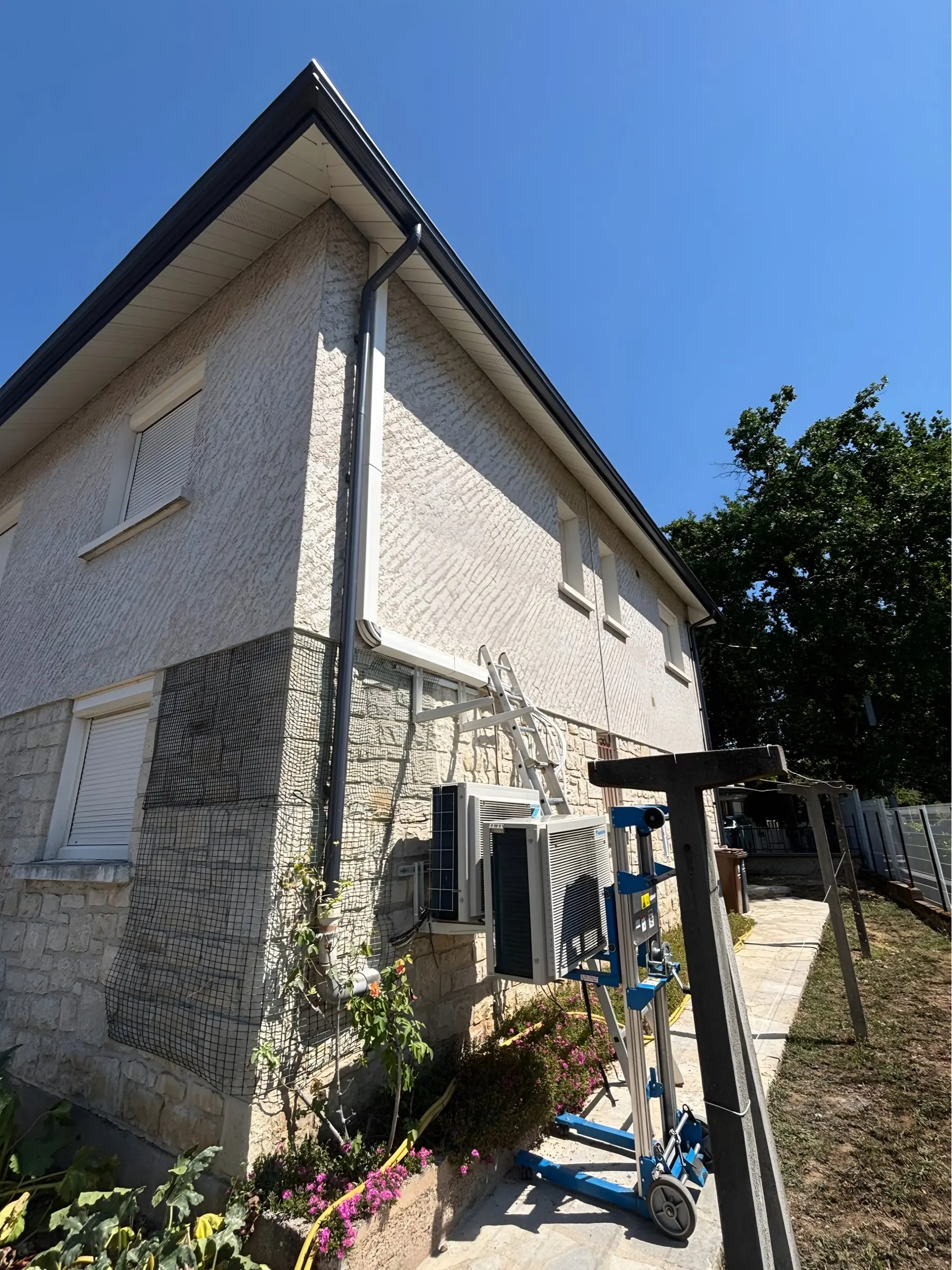 Façade crépie à soubassement de pierre : un groupe extérieur de climatisation sur un lève-charge bleu, à côté d’un autre déjà fixé sur ses équerres, une échelle contre le mur