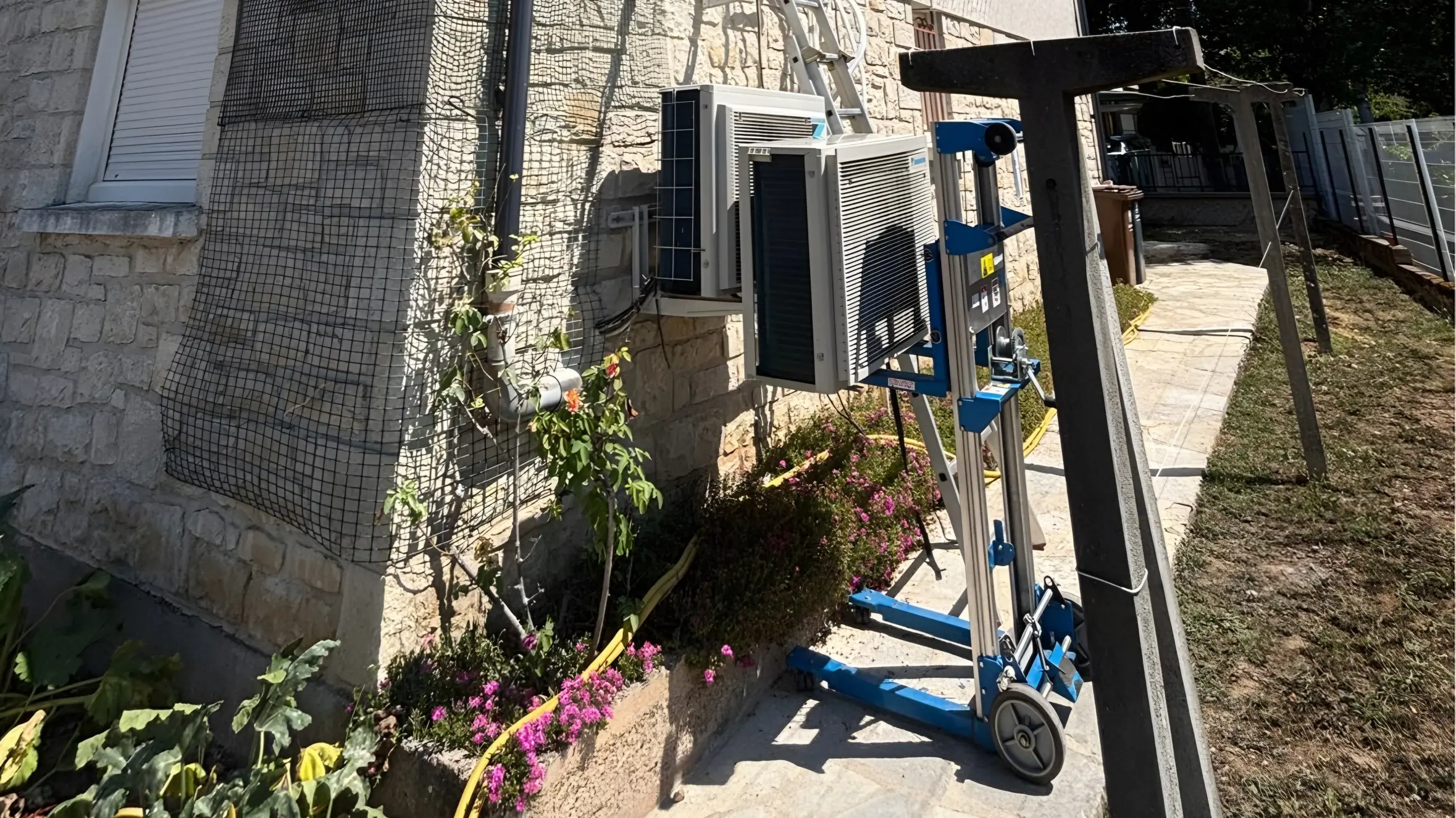 Façade crépie à soubassement de pierre : un groupe extérieur de climatisation sur un lève-charge bleu, à côté d’un autre déjà fixé sur ses équerres, une échelle contre le mur